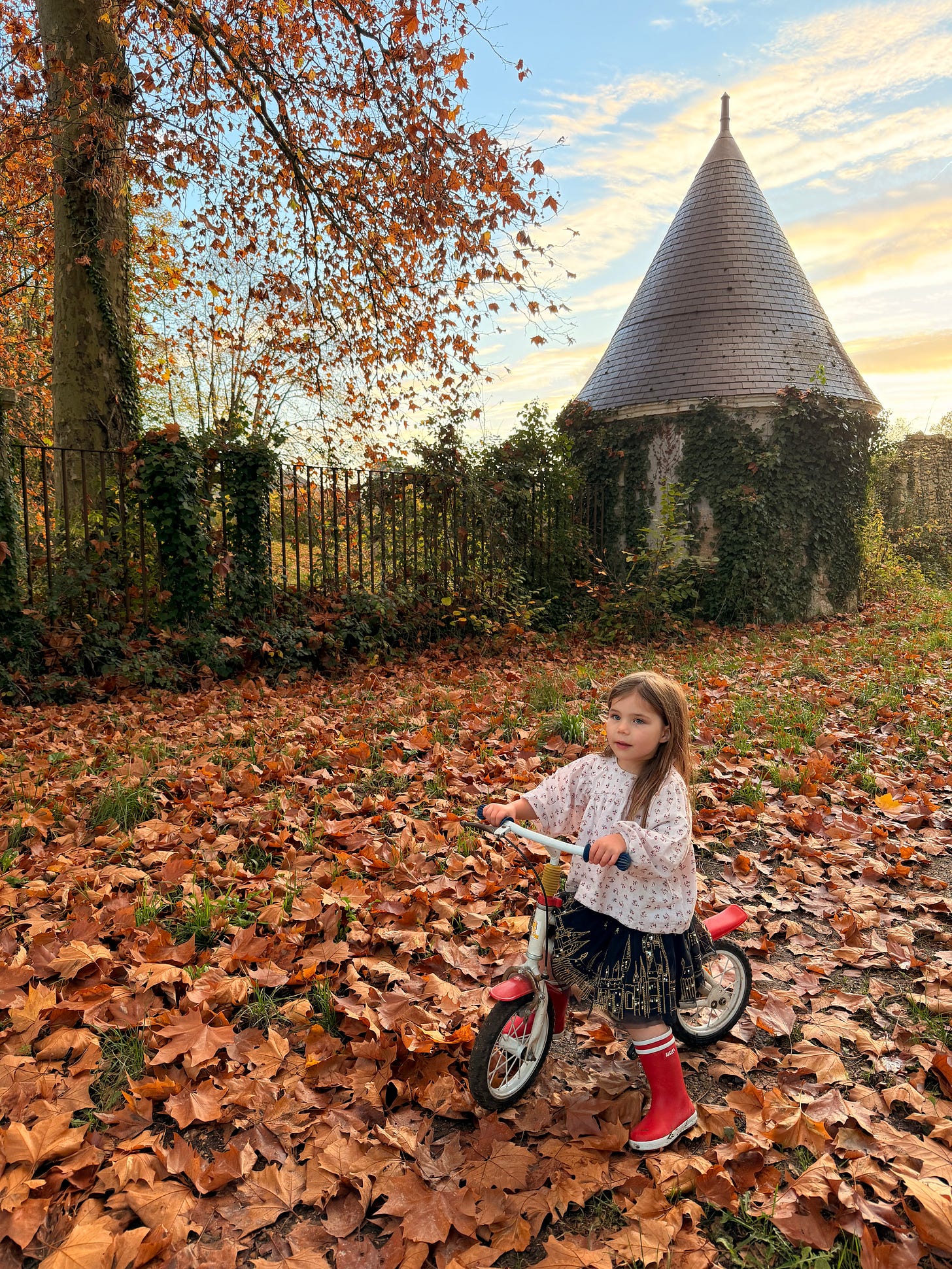 A young girl in a floral blouse, dark skirt and red rubber boots sits on a small red and white balance bike in a thick carpet of fallen leaves, with an iron fence and an ivy-covered round tower with a pointed slate roof behind her in golden evening light.