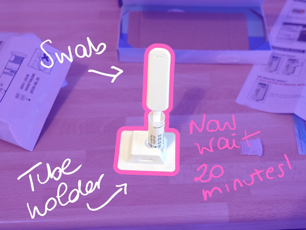 A close-up of an OraQuick HIV self-test in progress. The swab is inserted into the test tube, which is placed in a holder. The image has handwritten annotations indicating different parts of the test and a note stating, "Now wait 20 minutes."