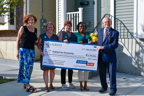 Photo of elected officials and Habitat homebuyers during a ribbon cutting, holding a large symbolic check from the City of Albany and a photo of staff from Community Preservation Corporation in front of Habitat homes.