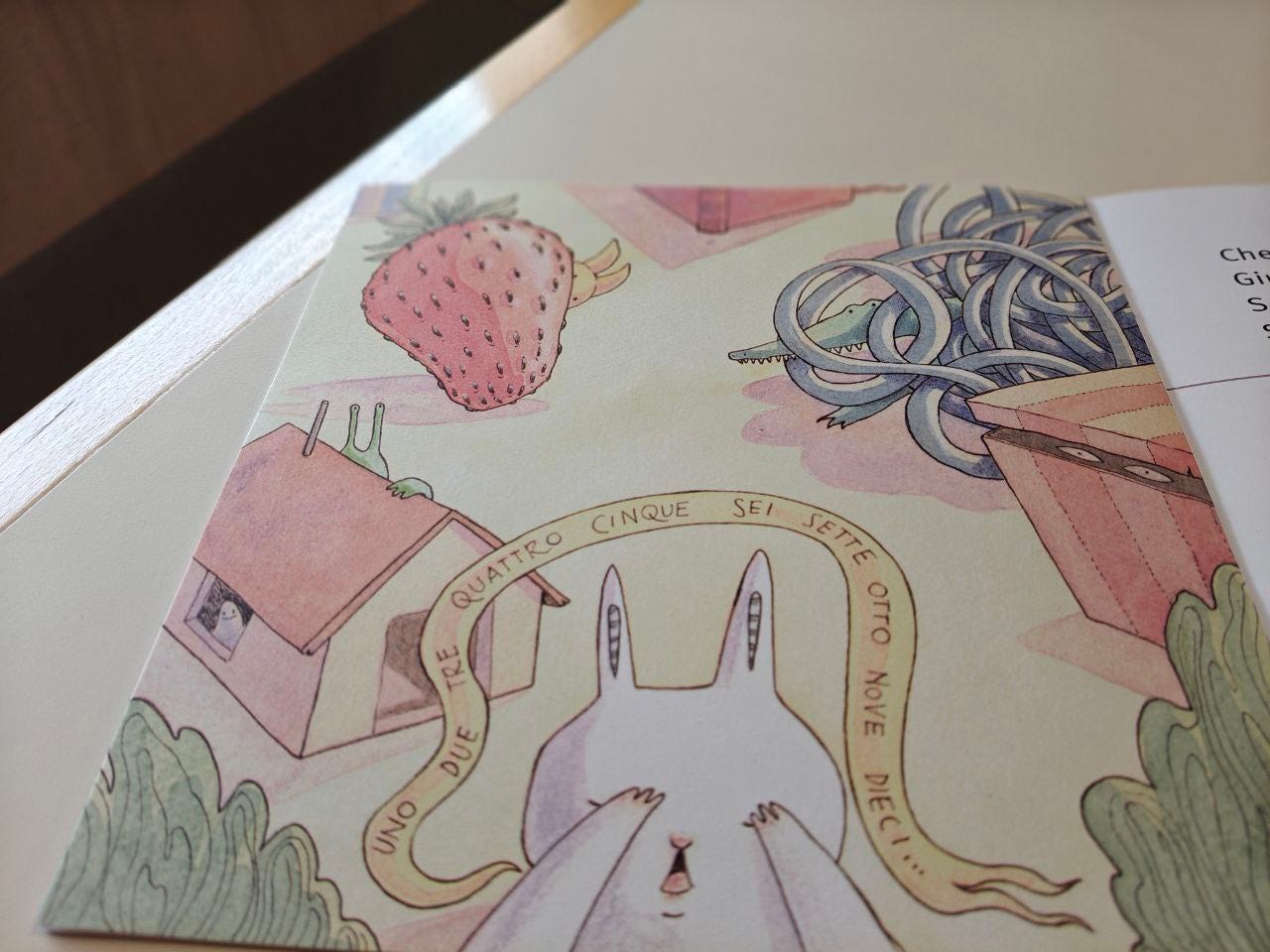 Una pagina della parola GIOCO delle Scatoline effequ con il disegno di un coniglio con le zampe sugli occhi nell'atto di contare, tanto che compaiono le parole UNO DUE TRE QUATTRO CINQUE SEI SETTE OTTO NOVE DIECI... Dietro di lui stanno nascosti: un fantasmino che sbircia dalla finestra di una casa, una creatura verde che sbuca con delle antenne verdi dal tetto della casetta, mezzo viso e due orecchie di una specie di lepre gialla, che sta dietro una fragola gigante, e un coccodrillo aggrovigliato dentro un tubo di gomma, e due occhi ch spiano da una scatola a righe arancioni appena socchiusa. Lo sfondo generale del disegno è giallo . La pagina è appoggiata su un tavolo di legno bianco sporco e nelll'angolino a sinistra un pezzo di muro.