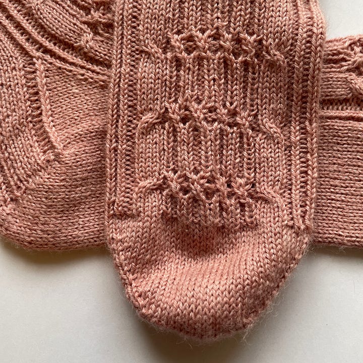 Two heavily cabled socks knit in pink yarn.