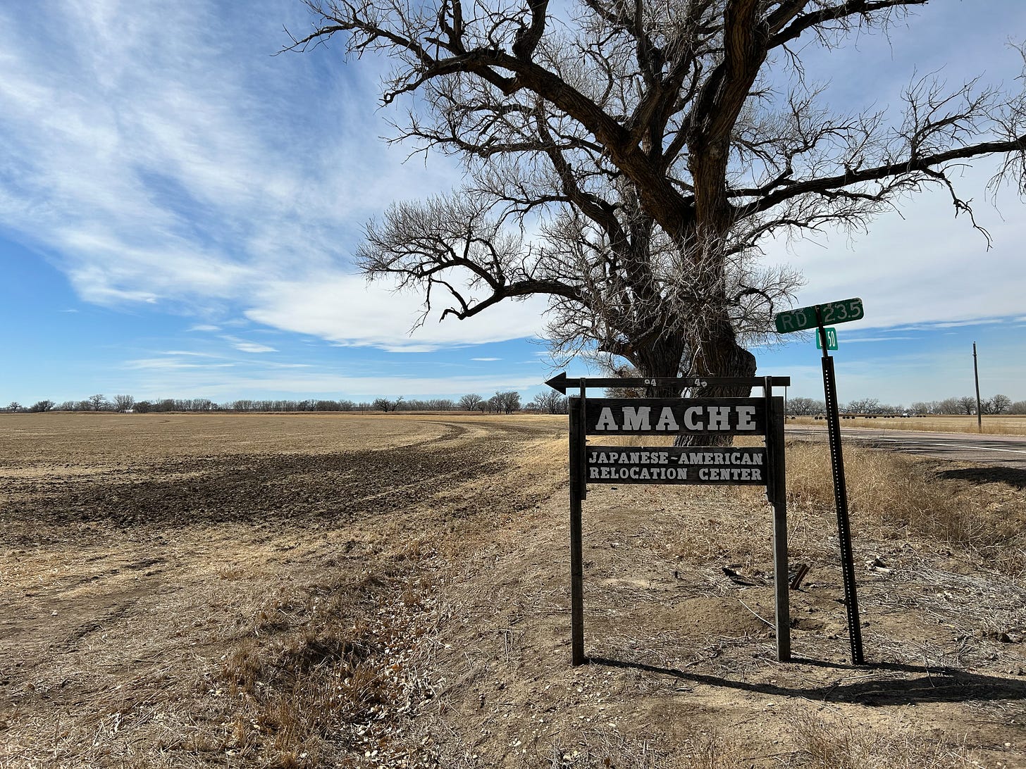 The entrance sign to the site that reads "Amache Japanese-American Relocation Center" in white lettering on a dark wooden sign. The background is an early spring bare agricultural field, a large tree without leaves, and a bright blue sky with an opaque blanket of soft and thin white clouds.