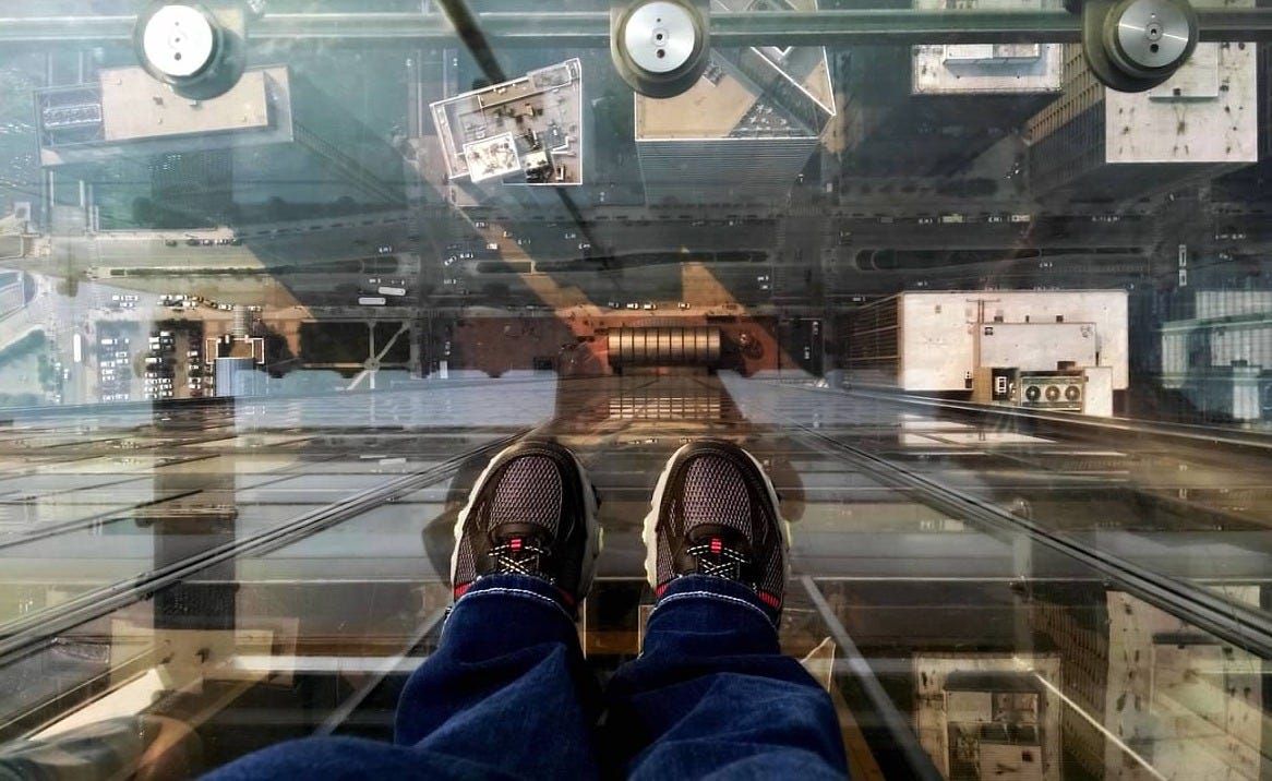 A few of two sneakered feet standing on a glass platform high above Chicago, looking straight down at the rooftops and streets below A few of two sneakered feet standing on a glass platform high above Chicago, looking straight down at the rooftops and streets below