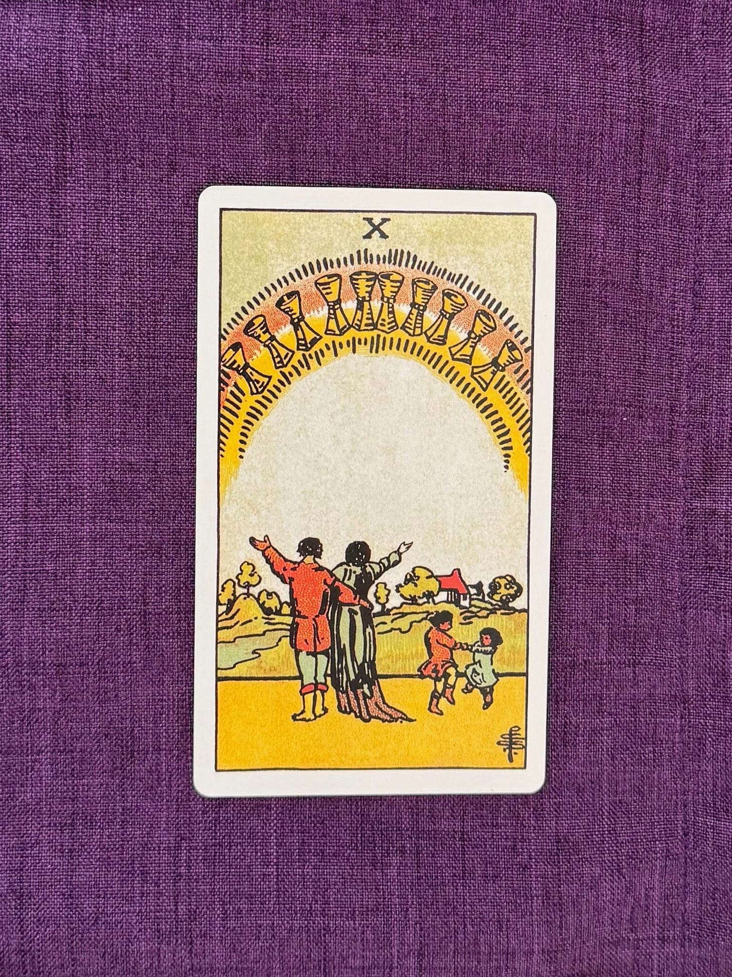 ten of cups Tarot card shows a family happily standing in front of the horizon. The parents face the children who are playing. There are ten cups on top, forming an arch.
