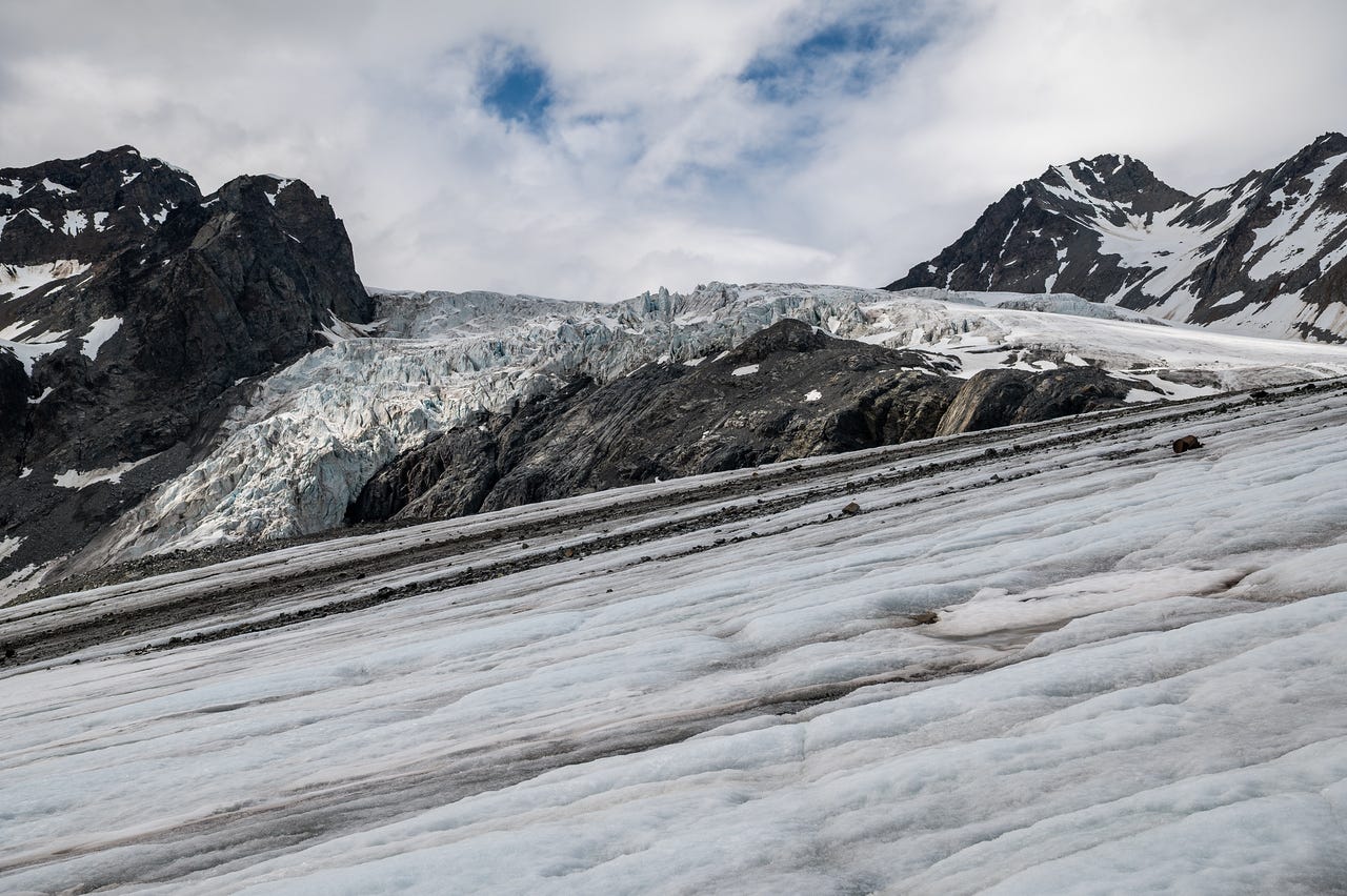 Looking up the white surface of a glacier toward an icefall, where the smooth ice buckles into a chaotic wall of pale blue seracs and crevasses spilling between dark rock buttresses. Bands of rock debris streak the ice in long diagonal lines. Snow-covered peaks flank both sides under a sky of thin cloud with one patch of blue. The foreground ice is rippled and streaked with meltwater channels.