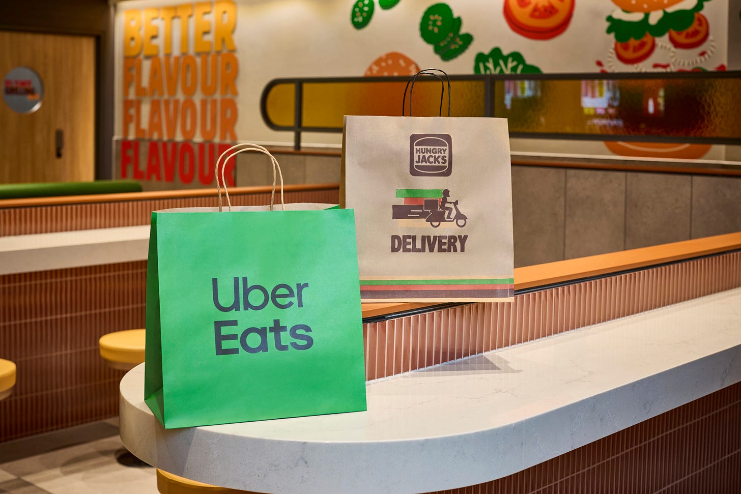 Green Uber Eats bag and brown Hungry Jack's delivery bag on a counter in a fast food restaurant with colorful wall art and seating in the background. Green Uber Eats bag and brown Hungry Jack's delivery bag on a counter in a fast food restaurant with colorful wall art and seating in the background.