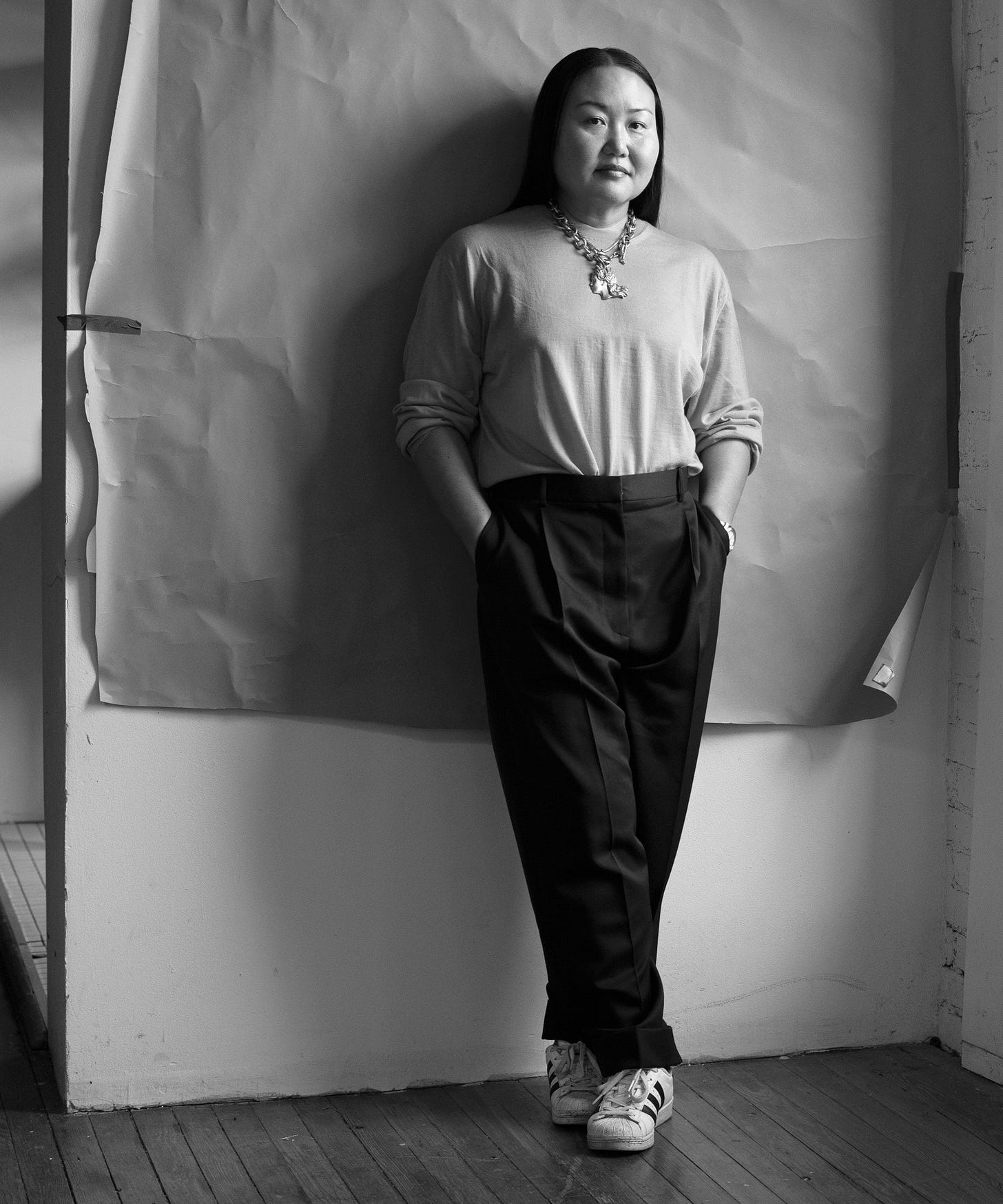 Hanya Yanagihara wearing a long sleeve tee chunk necklaces black slacks and Adidas shoes while leaning against a wall. Hanya Yanagihara wearing a long sleeve tee chunk necklaces black slacks and Adidas shoes while leaning against a wall.