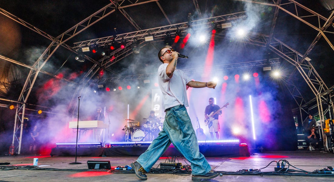 A man wearing a white t-shirt and jeans walking on stage with a band behind him A man wearing a white t-shirt and jeans walking on stage with a band behind him