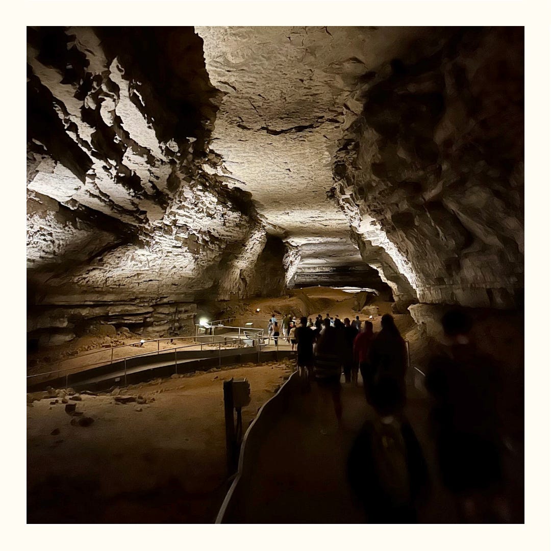 The dimly lit Mammoth Passage is visible, with a group of tour-takers experiencing the view. The cave is wide open, with boardwalk-style trails for tour-takers. The dimly lit Mammoth Passage is visible, with a group of tour-takers experiencing the view. The cave is wide open, with boardwalk-style trails for tour-takers.