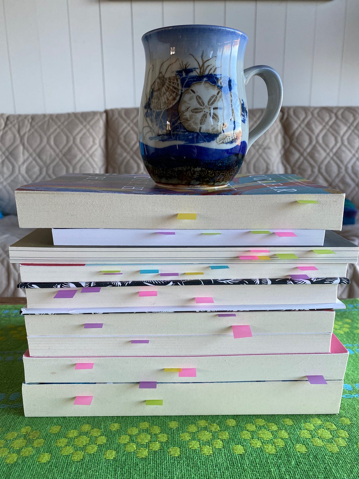 A stack of books, spines facing away. Small colored post-it notes poke out from the pages. A ceramic mug painted with a sand dollar sits on top of the books.