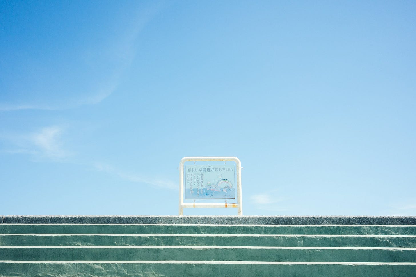Sfondo di cielo azzurro che occupa i tre quarti superiori della foto, mentre sotto c'è una scalinata di cinque gradini verde smeraldo di cemento, e in cima un cartello alto mezzo metro con delle scritte in caratteri giapponesi.