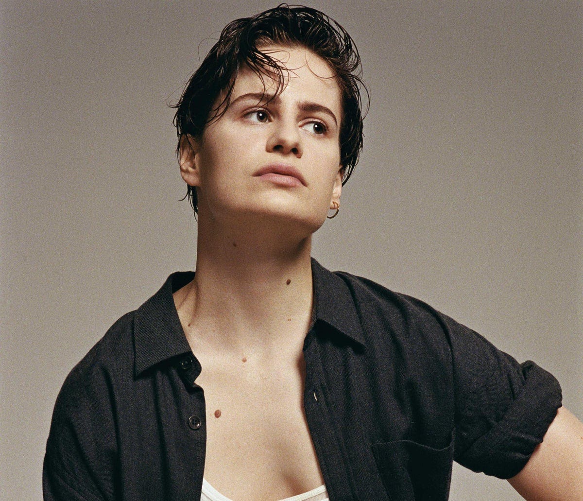 A photograph of a genderqueer person wearing an open button down shirt and staring into the middle distance.