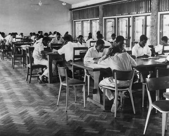 A black and white photograph of people sitting at long tables, reading A black and white photograph of people sitting at long tables, reading
