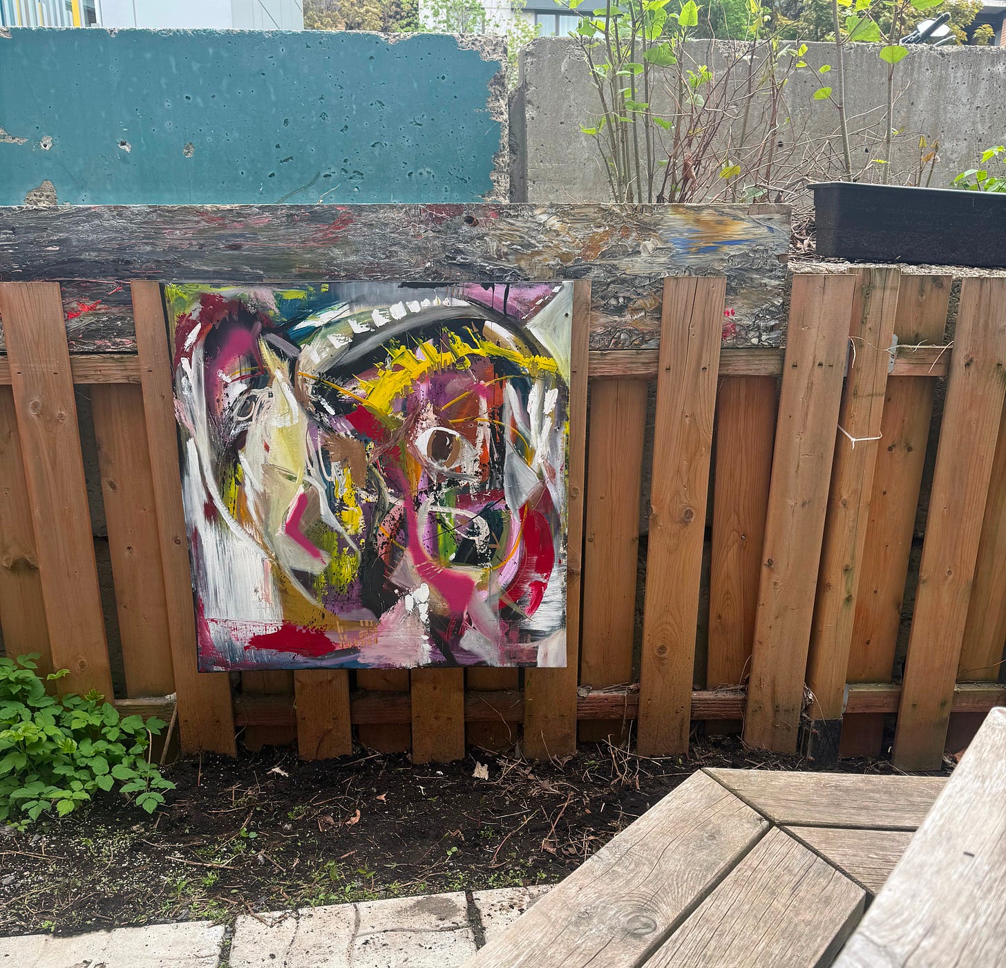An abstract painting screwed to a wooden fence in a backyard. A weathered picnic table sits in the foreground. Concrete blocks and early spring growth are visible beyond the fence.