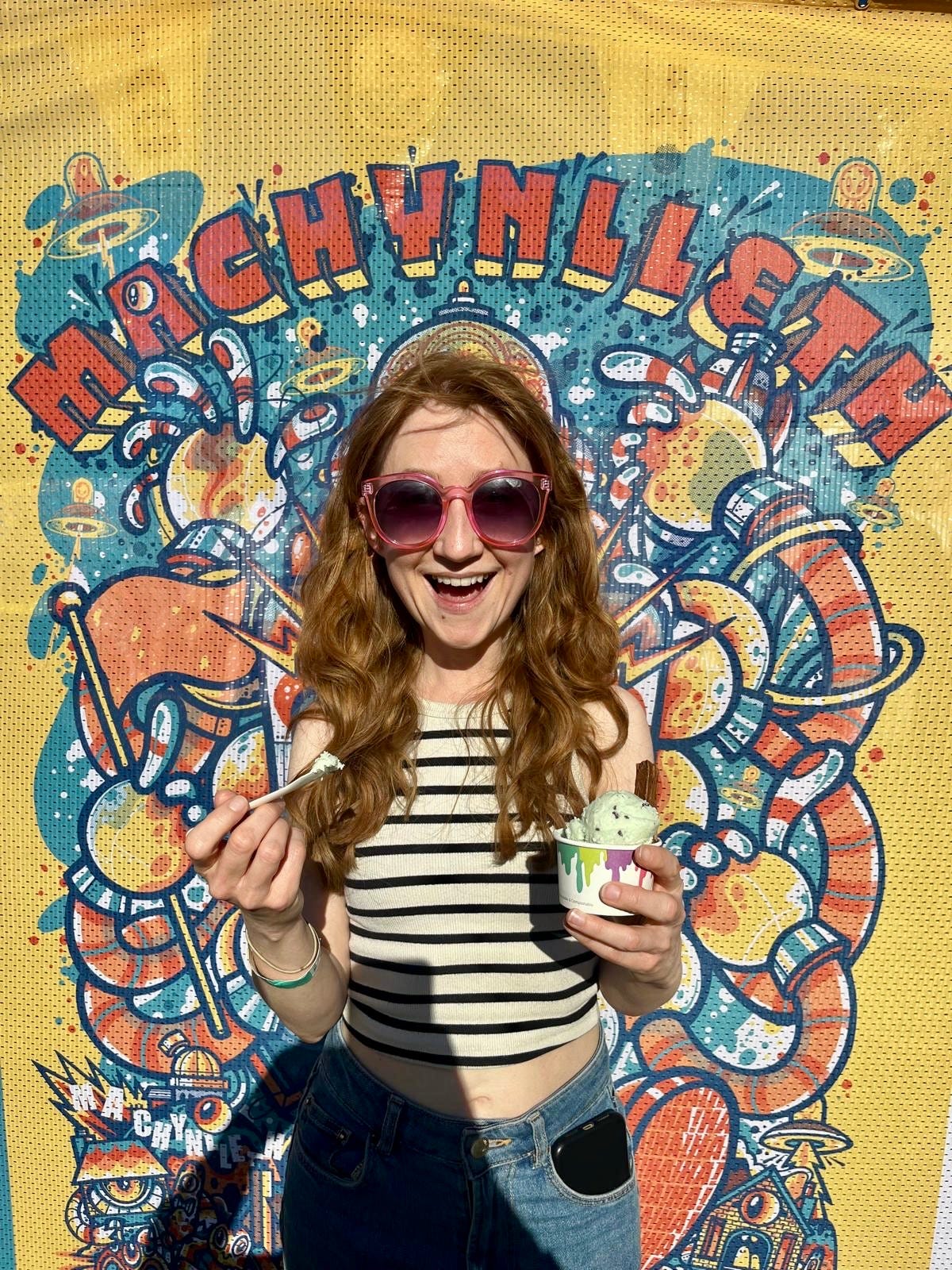 Woman with long ginger hair holds an ice cream and smiles at the camera. Macf Fest logo behind her Woman with long ginger hair holds an ice cream and smiles at the camera. Macf Fest logo behind her
