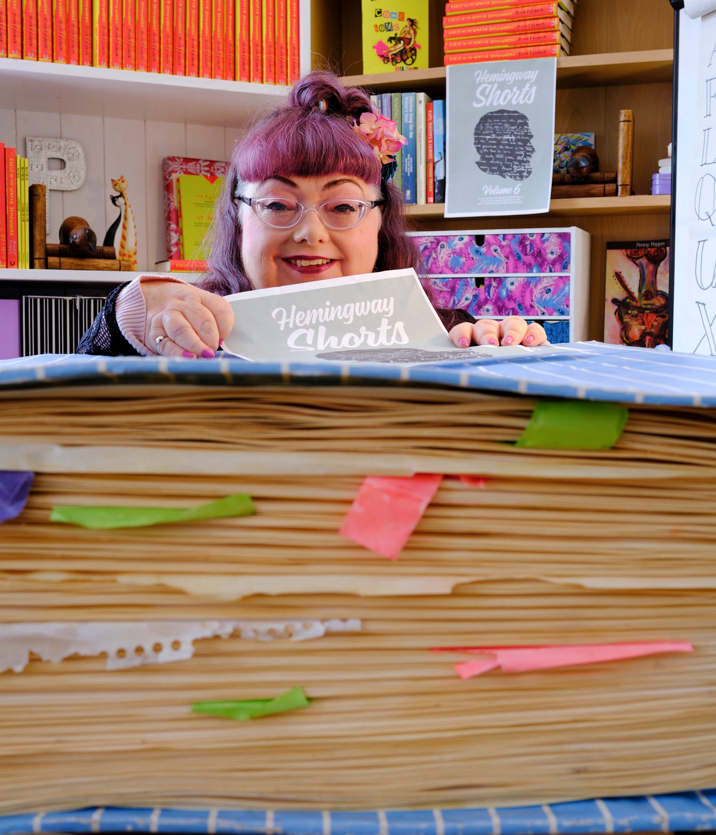 Penny sits in front of bookcases which show off her published work. First in the World Somewhere is a red book with yellow writing. Come Home Alive is a yellow book with multiple-coloured typeface and a picture of Penny in a steampunk wheelchair. There is also an image of Hemingway Shorts Volume 6 behind Penny's head and a flyer she holds in her hand. At the front of the image is a large replica of one of her diaries used in her one-woman show, Lost in Spaces. Penny has pink red hair, a pink flower and wears glasses. The whole image is colourful and lively.