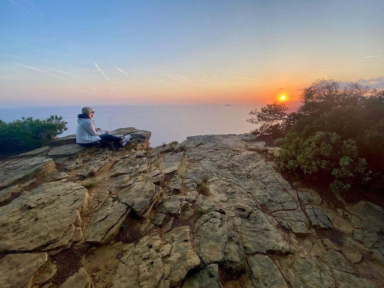 Kelly sits alone on a rocky overlook at sunset, facing the sea and orange sky. Kelly sits alone on a rocky overlook at sunset, facing the sea and orange sky.