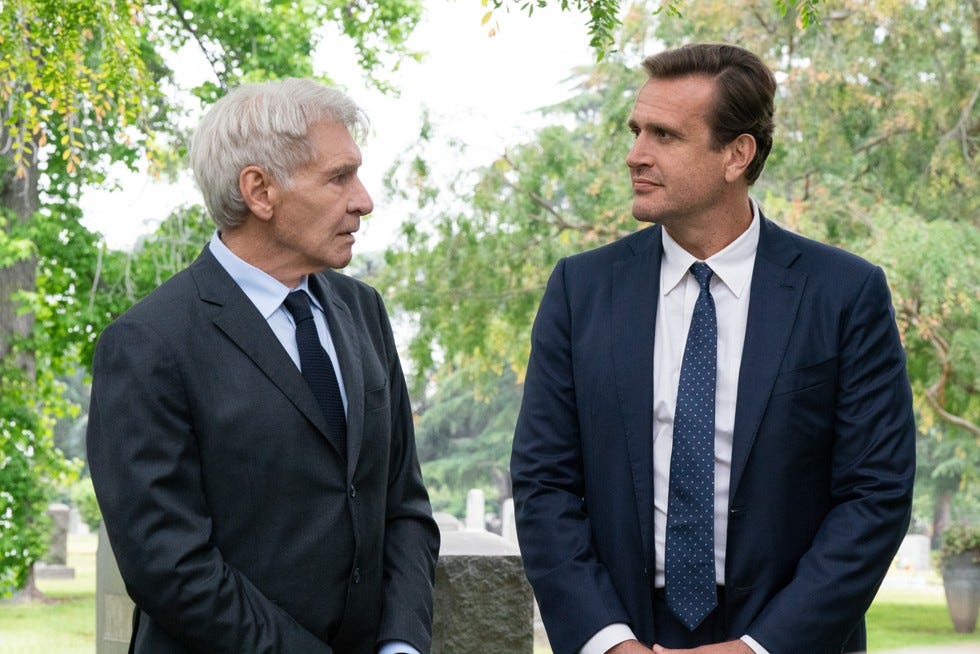 Jason Segel and Harrison Ford in Shrinking, both wearing black suits and ties, stood together outside.