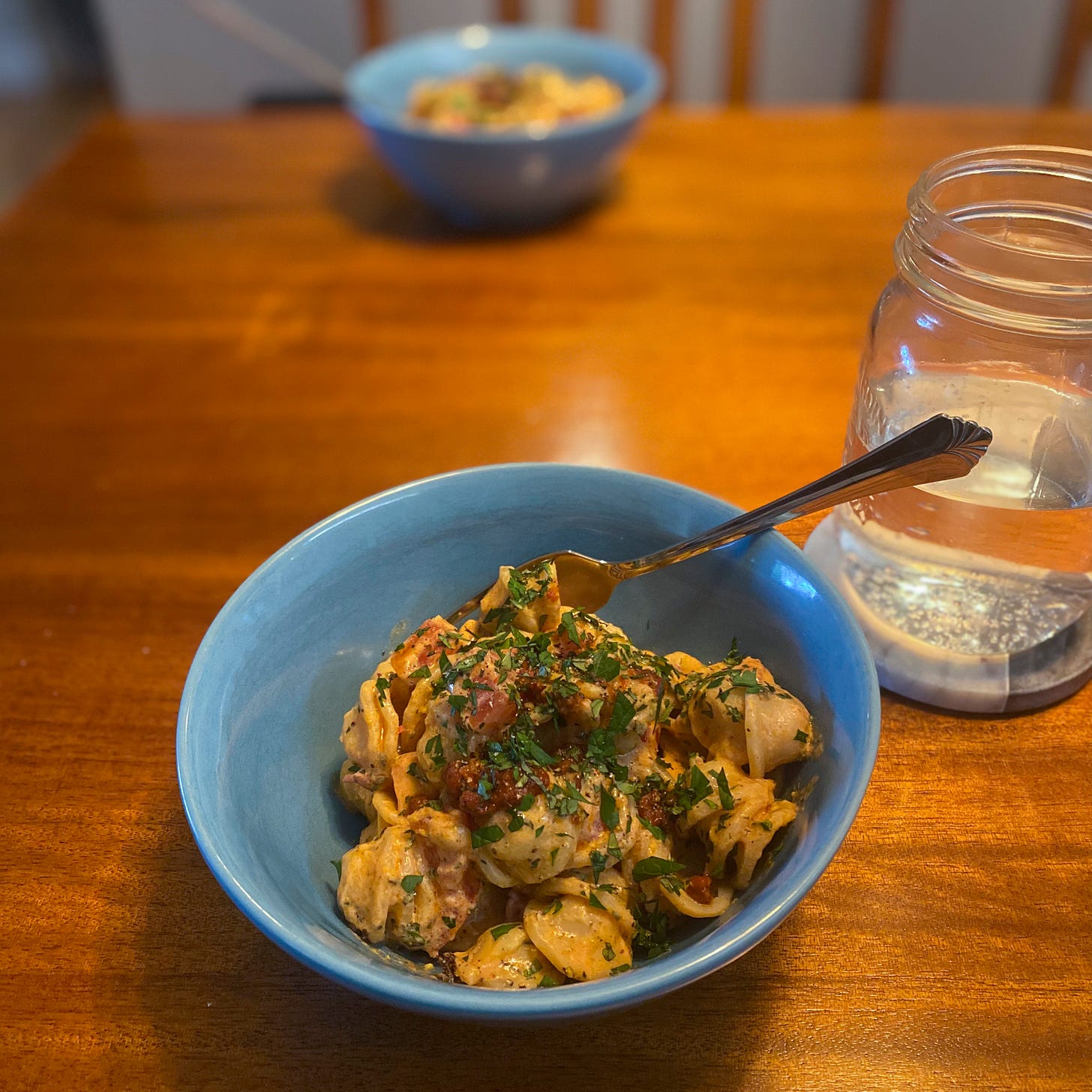 Blue bowls across from each other on a wood table. Each is filled with orecchiette in an orange-ish sauce, and topped with herbs and extra of the tomato paste-brown butter mixture. A glass of water sits on a coaster beside the bowl in the foreground.