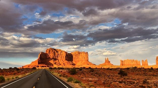 A road in the desert. On the horizon are typical American towering rock formations resembling those of Monument Valley that rise dramatically from the desert floor. I took this photo in the late afternoon, so the setting sun gives the rocks a warm glow, characteristic of the ‘golden hour’.