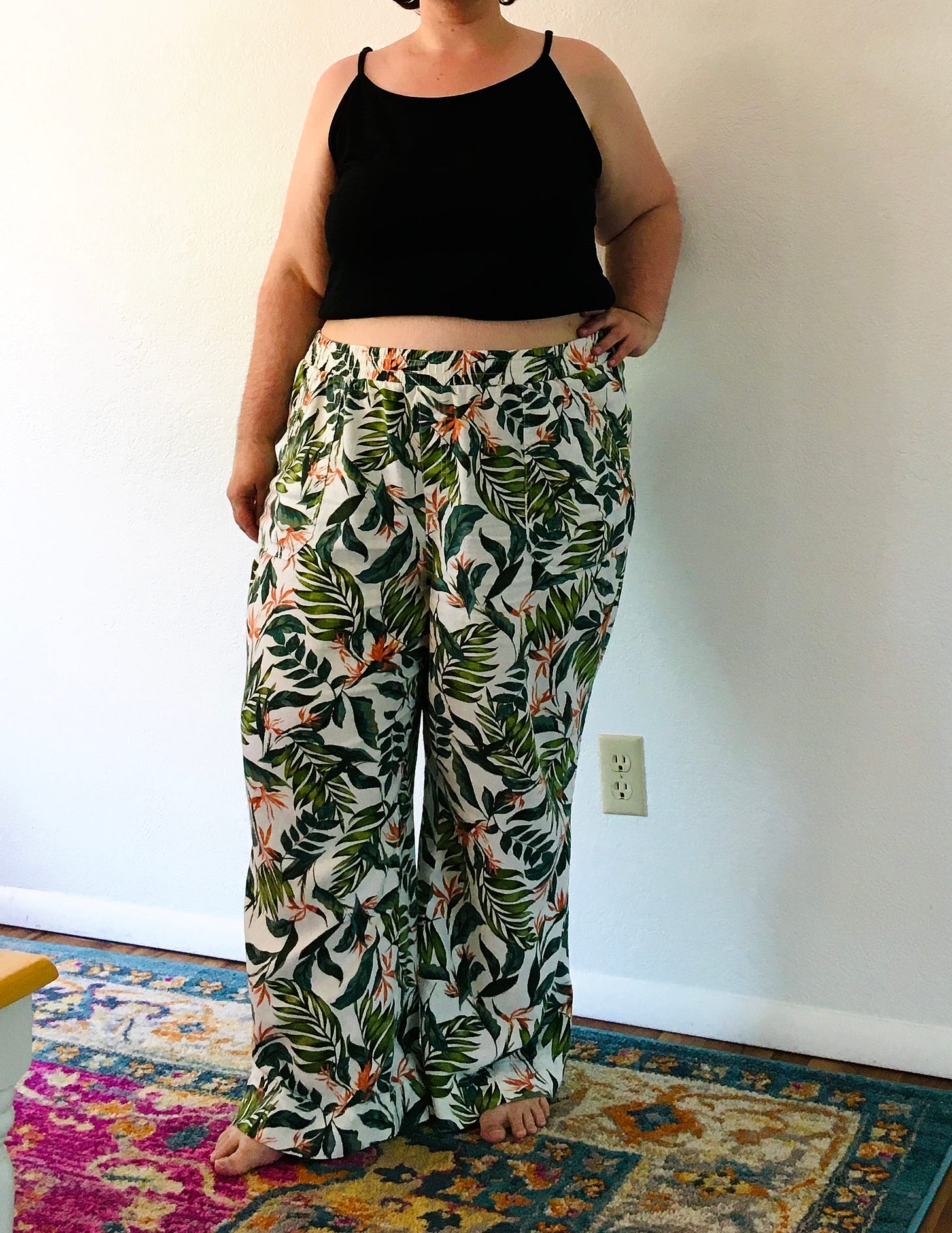 A fat white fem stands in front of a white wall. She’s wearing a black cropped cami and pants that are cream colored with large palm fronds and bird of paradise flowers scrattered across them
