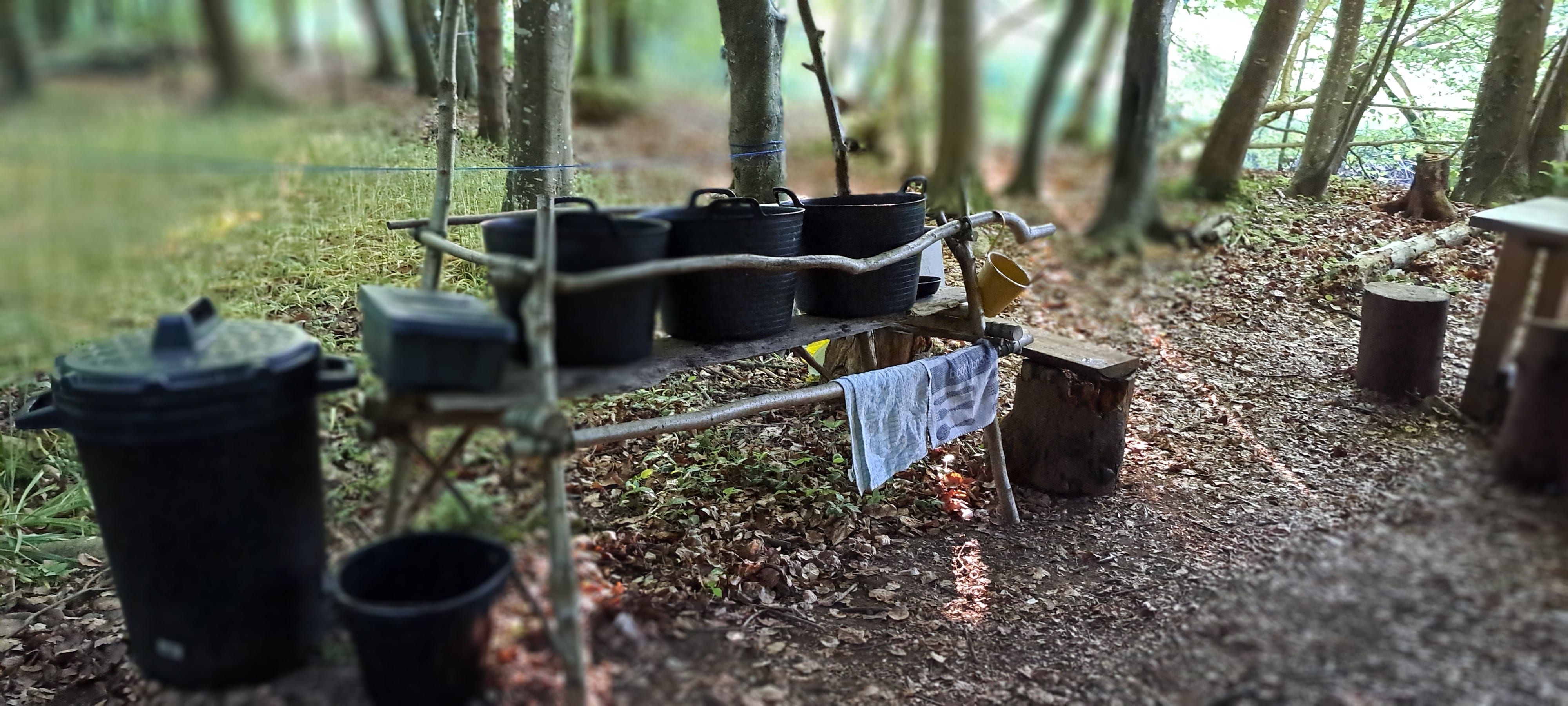 The washing op station consists of a bench made of planjks and tree branches into which 3 large black tubs are sitting. Tea-towels are hanging over a rail made from a tree branch. A smaller bench on the left is where clean crockery is left to dry. To the right is the dining area, just seen at the edge of the image. 