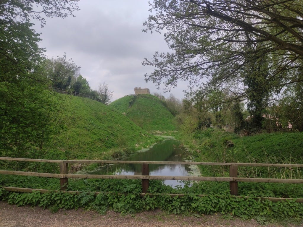 Wooden railings overlooking a stream (which used to be part of the moat).  Behind is a mound a few metres high, on top of which runs the "Lady's walk", and then another, higher mound which holds the remains of the castle keep.