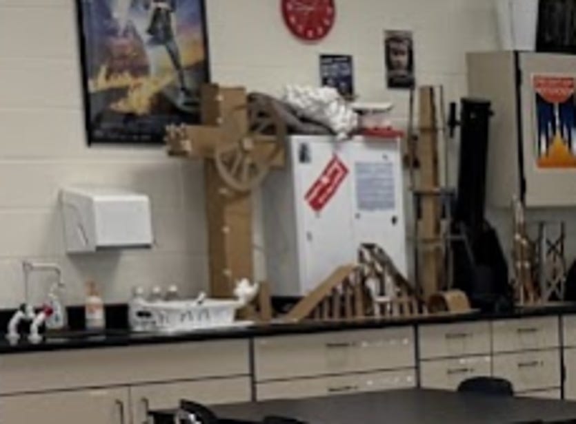 A physics classroom display featuring several student-built Bad Idea Park ride models arranged along the back counter. The largest model appears to be Jesus Take the Wheel—a towering cardboard cross with a Ferris wheel attached near the top—surrounded by other wooden and cardboard amusement ride prototypes. The collection showcases the final engineering projects created by students as part of a physics design challenge. A physics classroom display featuring several student-built Bad Idea Park ride models arranged along the back counter. The largest model appears to be Jesus Take the Wheel—a towering cardboard cross with a Ferris wheel attached near the top—surrounded by other wooden and cardboard amusement ride prototypes. The collection showcases the final engineering projects created by students as part of a physics design challenge.