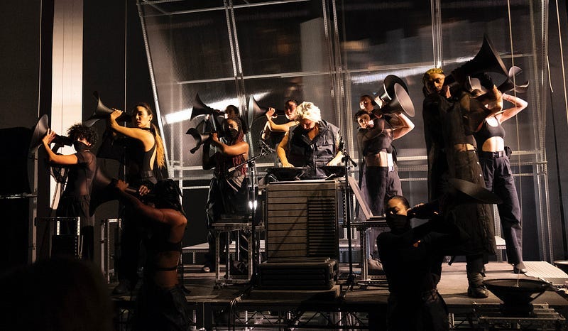 Performers dressed in all-black face front, standing on a stage. The setting is stark and industrial: concrete columns flank the dimly lit view and the bare-bones stage makes no effort to conceal the scaffolding underneath. There’s a textured backdrop of ribbed plexiglass. The performers hold black megaphones up to their ears.