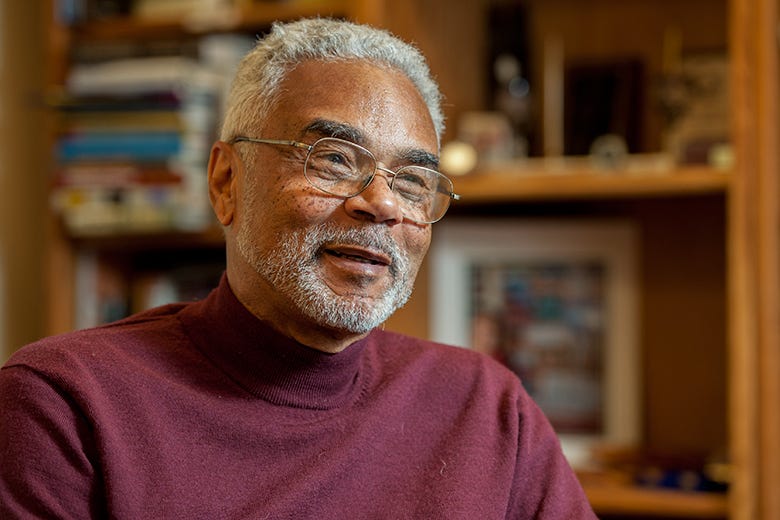  Dr Carson, an African American man with wire rim glasses, white hair, and a close cut white beard, wears a brown turtleneck sweater. He is smiling and looks like he is in mid-conversation. Behind him are bookshelves at the King Institute.