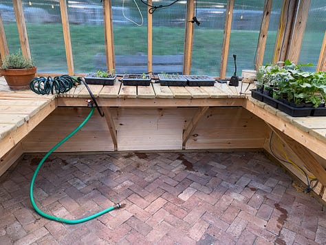 Alt text: Three side-by-side photos of a greenhouse interior during construction. The images show newly built wooden raised planting beds arranged around the perimeter of the space, with a herringbone-pattern brick floor installed throughout the center walkway. The greenhouse framing and clear wall panels are visible, and the beds are unfinished, showing the structural supports beneath the bench tops. The photos capture different angles of the same space, highlighting the layout of the raised beds and brick flooring.
