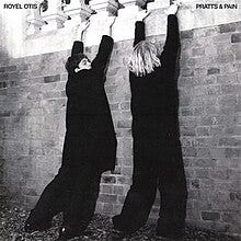 The two band members, wearing baggy all-black clothes, their backs to the camera, lean up against a brick wall and hold onto the balcony above them.