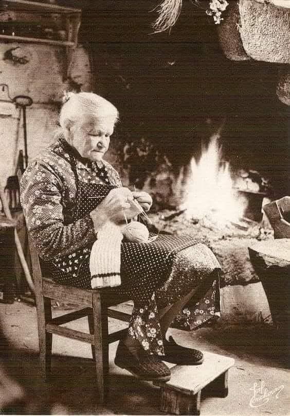 This may contain: an old woman knitting in front of a fire