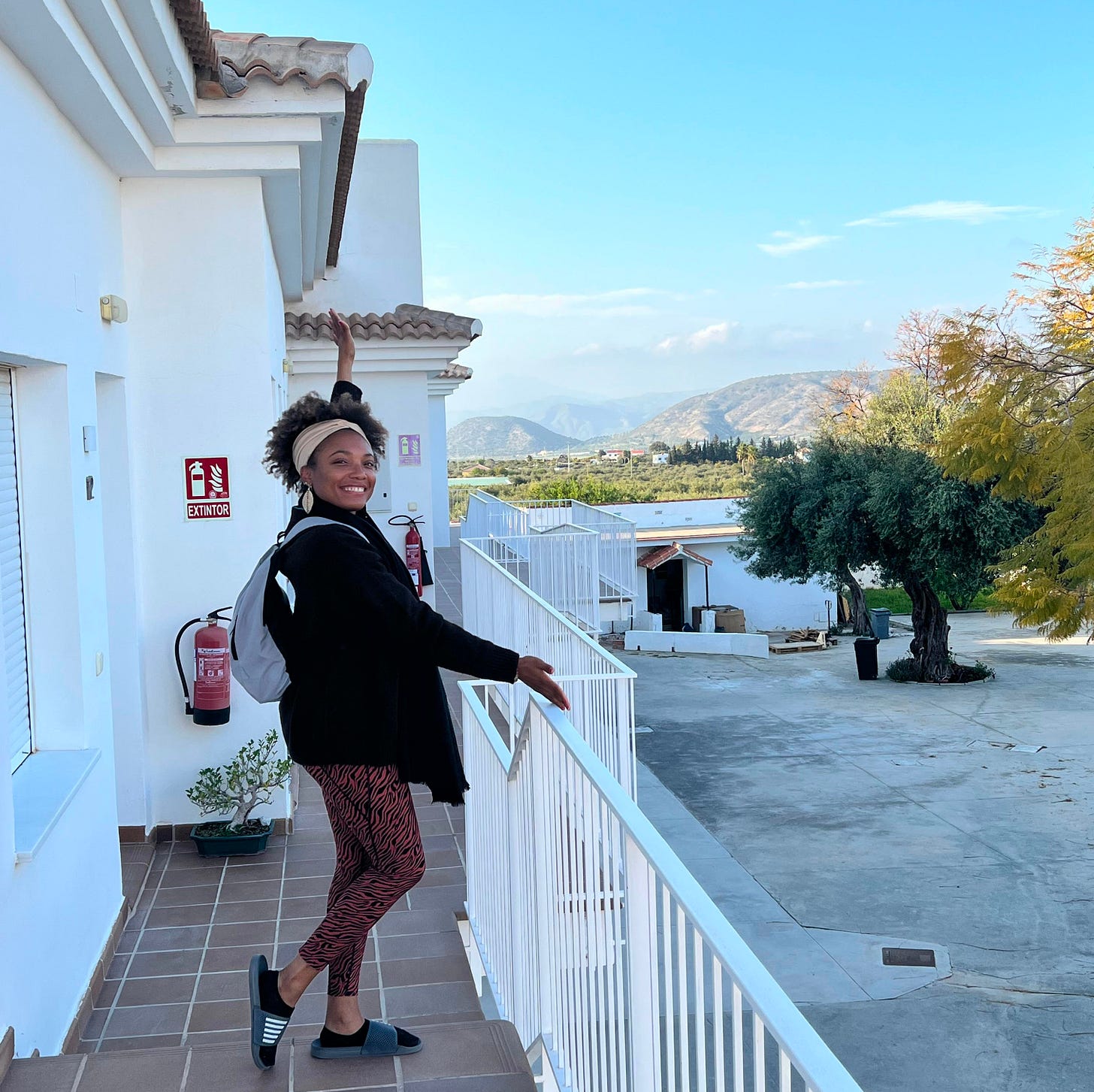 The author is in orange zebra print leggings, posing playfully on a white Andalusian building balcony with mountains and olive groves visible behind her. The author is in orange zebra print leggings, posing playfully on a white Andalusian building balcony with mountains and olive groves visible behind her.