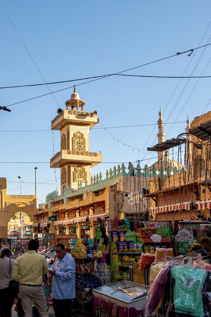 Top left: A busy market street in Aswan with vendors and shoppers beneath a mosque minaret and tangled overhead wires Top right: Two women sitting together sharing a phone beside a table of cosmetics and bottled goods, with a woman in a niqab seated nearby Bottom right: An elevated view across the rooftops of old Aswan toward the Nile, with feluccas and cruise ships moored along the banks and the modern city beyond Bottom left: A young man crouched over an open telecommunications junction box with tangled colored wires, while a woman in a pink hijab and a shopkeeper stand in the doorway behind him