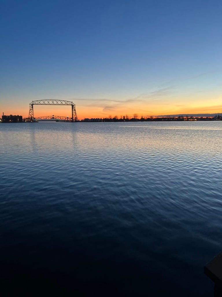 Duluth's arial lift bridge sits at the mouth of the St. Louis river where it flows out of Lake Superior. A band of orange along the horizon line fades into blue as the sun rises.