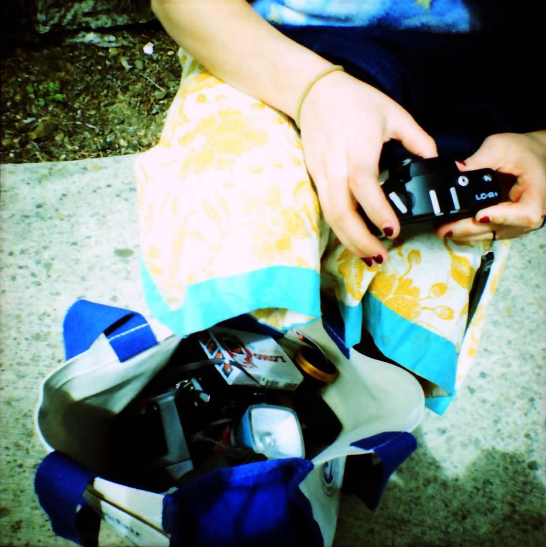 A saturated close-up photo of Sarah's arms and hands. She holds a film camera, and a bad of film accessories sits on the ground in front of her. She seems to be outside on sidewalk, and wearing a yellow and blue skirt.