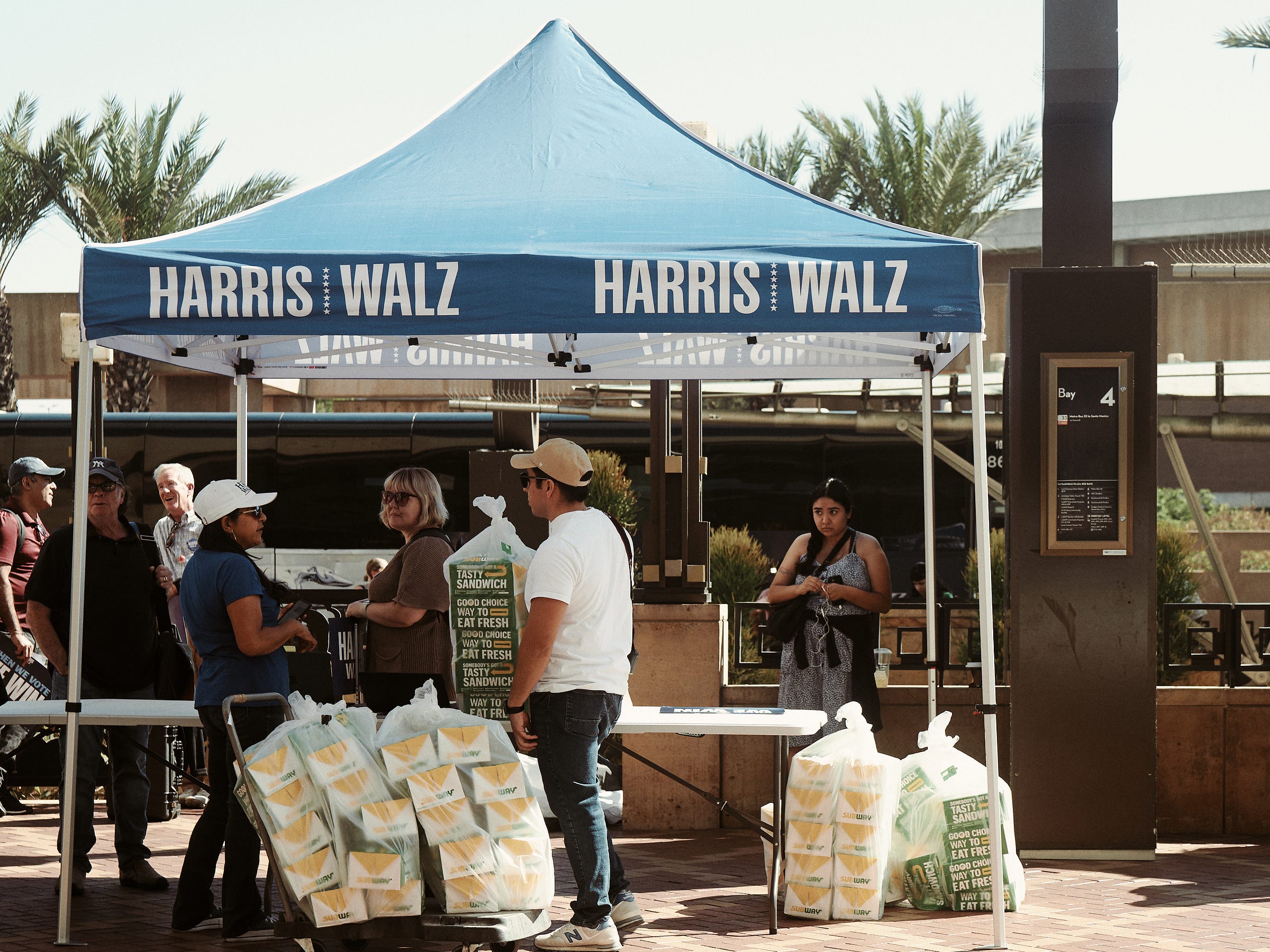 People gather under a shaded Harris Walz cover as they setup the check-in process for the bus ride to Phoenix, AZ. Photographed by LA photographer Afonso Salcedo.