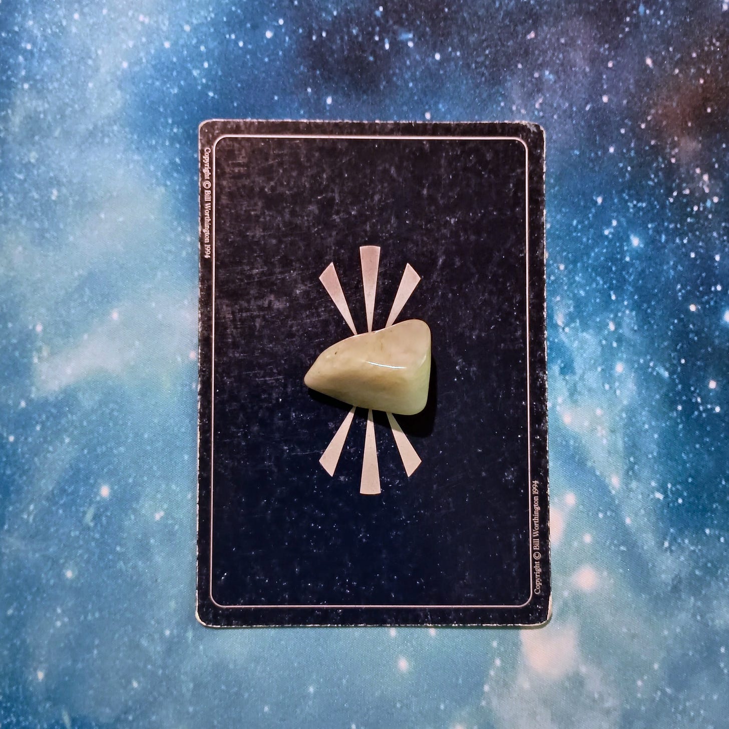 New Moon Magic: Your July 2025 Pick-a-Card Tarot Reading, pile 3 - Jade (light green) stone and an oracle card, face down, on a starry blue background