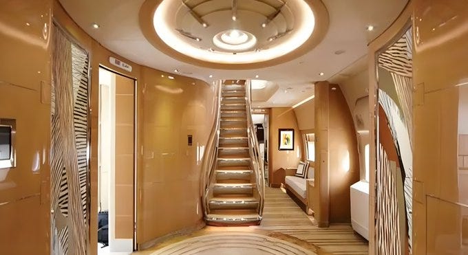 A luxurious interior of a private jet with polished wood paneling and metallic accents. A curved staircase with gold railings leads to an upper level. The hallway features sleek, modern decor with beige walls, intricate patterns, and recessed lighting. A bedroom area with a bed and artwork is visible in the background.