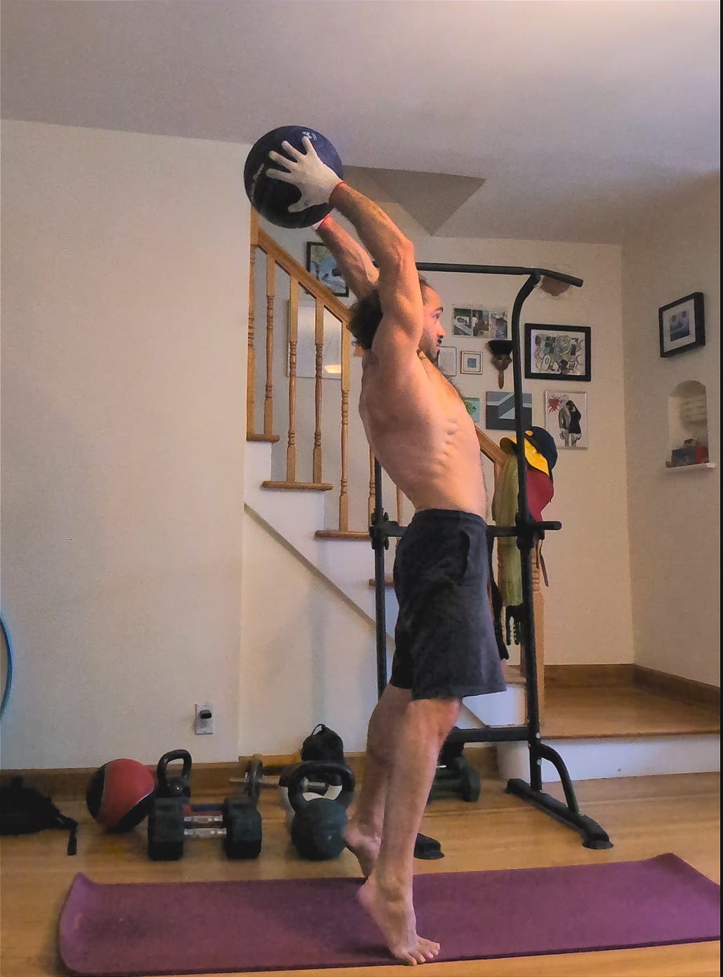 A man standing on his toes on an exercise mat with arms extended overhead, holding a slam ball in the process of slamming it down onto the mat.