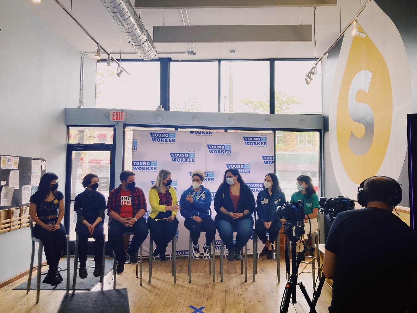 a panel of 8 people sitting in high top chairs pass the microphone to each other in front of a banner reading "young worker forum afl-cio" a panel of 8 people sitting in high top chairs pass the microphone to each other in front of a banner reading "young worker forum afl-cio"