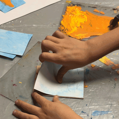 Child's hand pressing inked foam block onto blue paper Hands rolling orange ink onto a brayer Child rolling brayer over foam printing block Hand holding carved foam block with orange ink