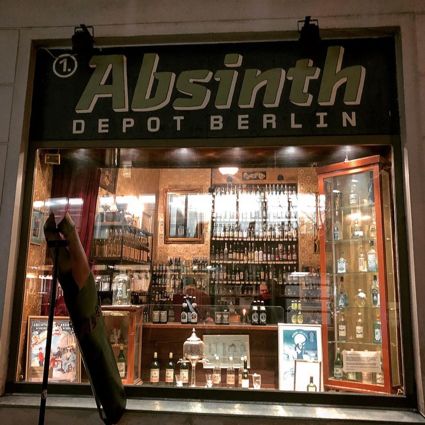 Shopfront of “Absinth Depot Berlin” at night, with illuminated shelves of bottles, vintage posters, and glassware visible through the window.
