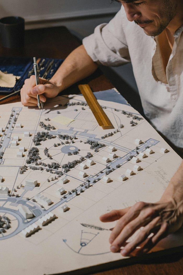 A man holding a pen and working on a scale model of an architectural plan, with tiny trees and sketches on the paper.