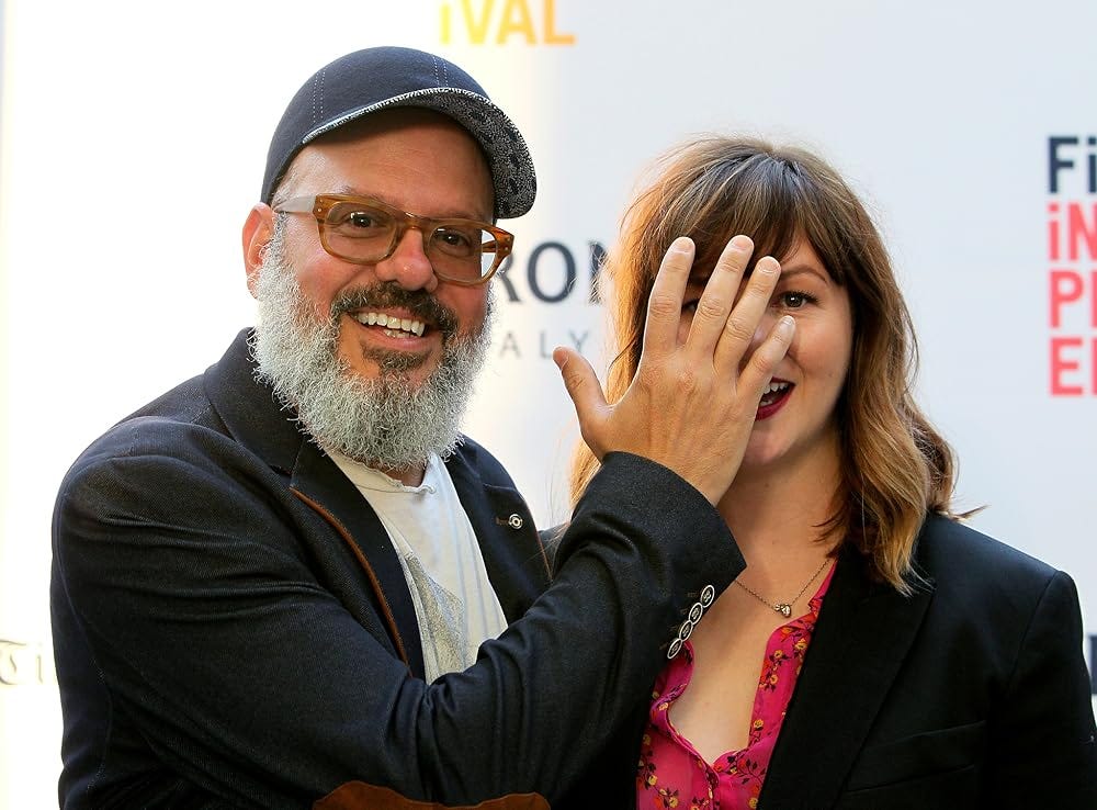 Amber and David pose together in front of a step-and-repeat. David smiles broadly while his hand blocks Amber’s face.