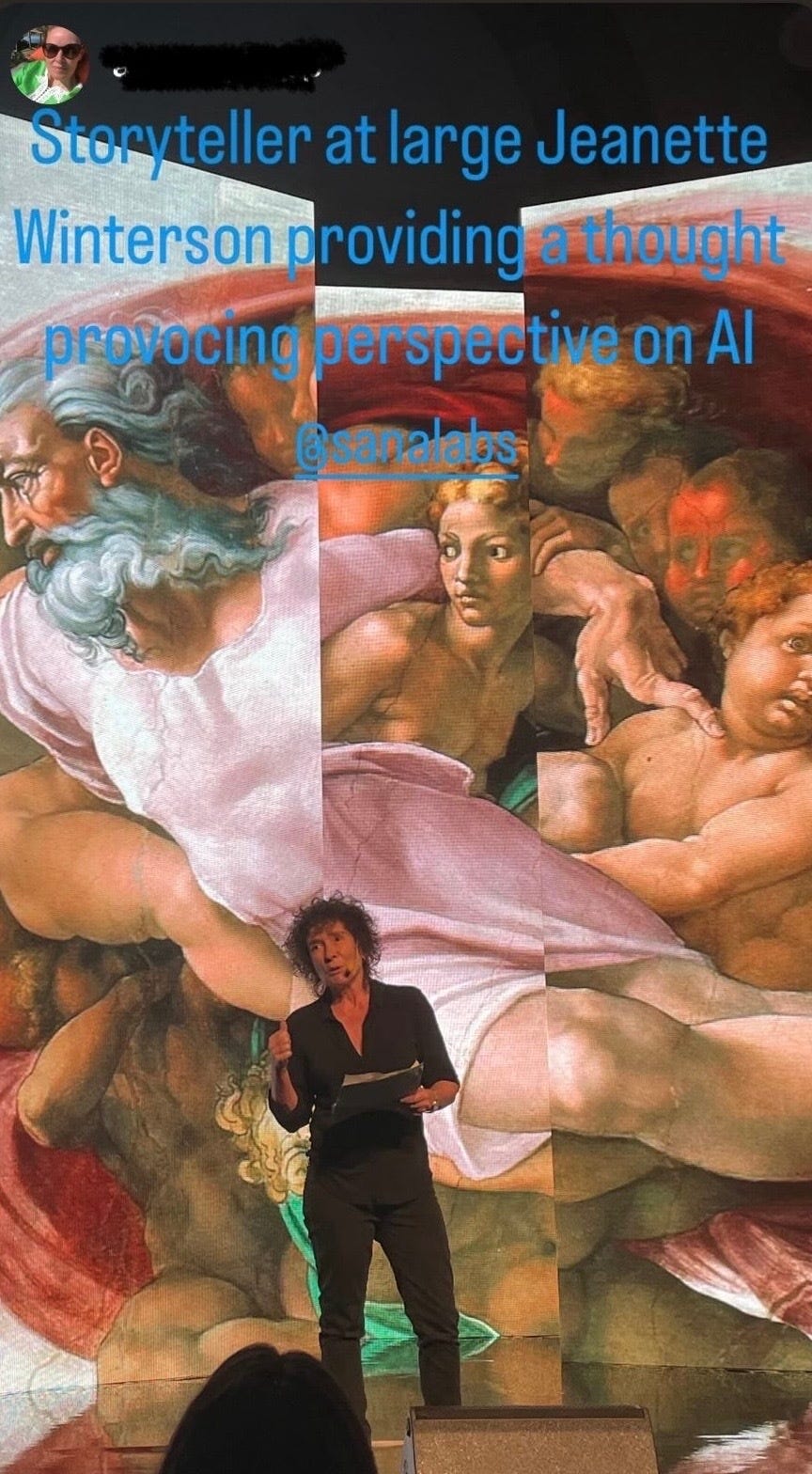 Photograph of Jeanette Winterson standing in front of tree LED screens showing Michaelangelo's Creation of Adam.