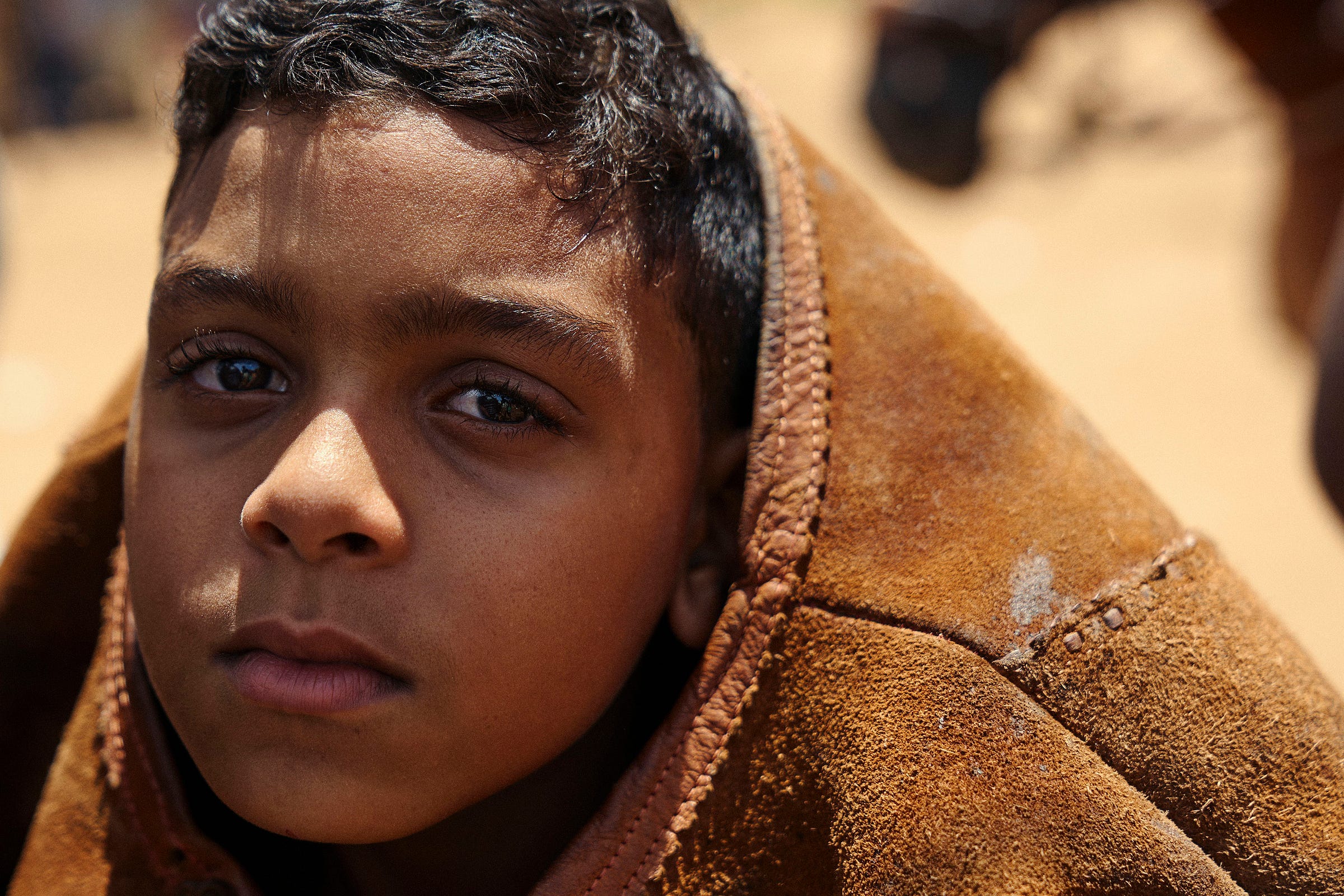 A young boy with short dark hair looks into the camera, wrapped in a brown, worn blanket or cloak. The background is blurred and sunlit.