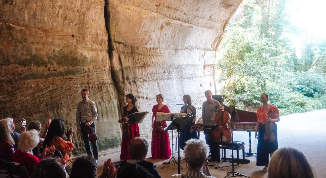 Six musicians stood in the entryway of a cave with an audience in front of them Six musicians stood in the entryway of a cave with an audience in front of them
