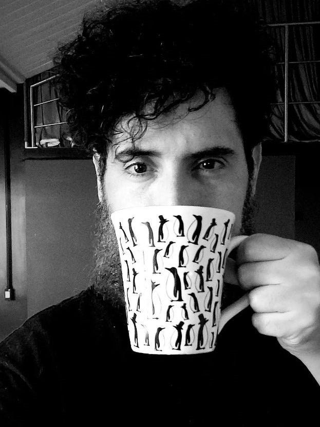 Imagem em preto e branco. O autor Eric Novello olhando diretamente para a câmera com uma caneca na frente do rosto cobrindo a boca e parte do nariz.
